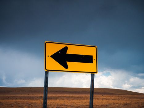 A bold yellow arrow sign against a dramatic overcast sky in a vast empty plateau landscape.